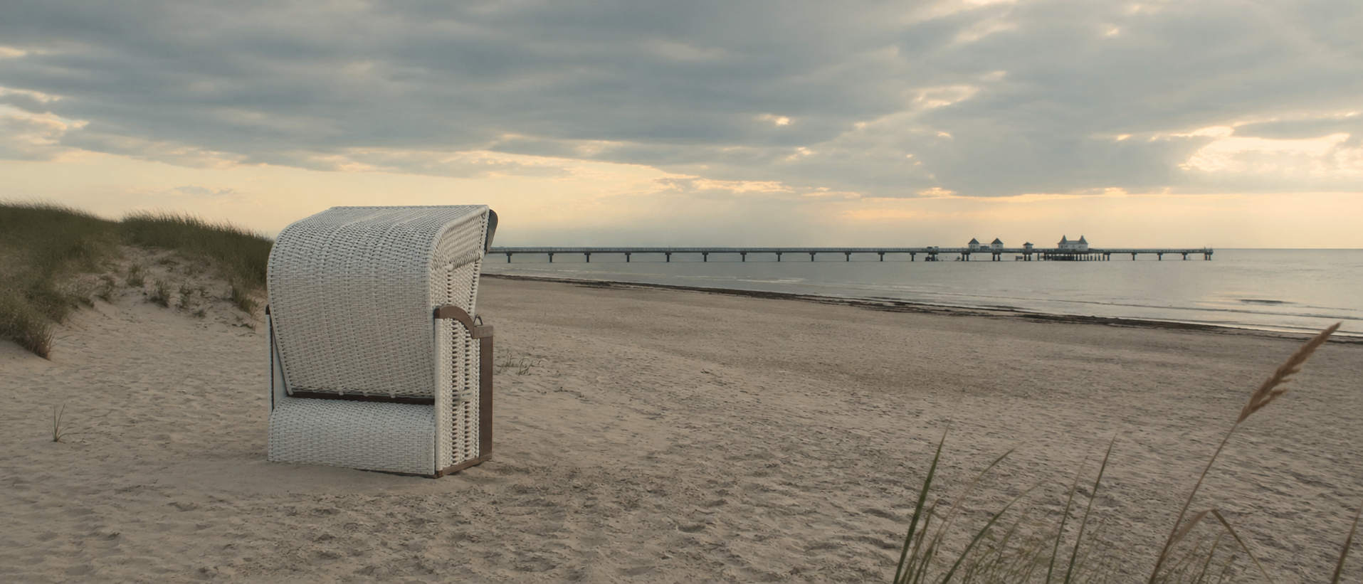 Weißer Strandkorb steht am Ostseestrand, Seebrücke im Hintergrund; Favorent kümmert sich um die Vermietung von Ferienimmobilien.