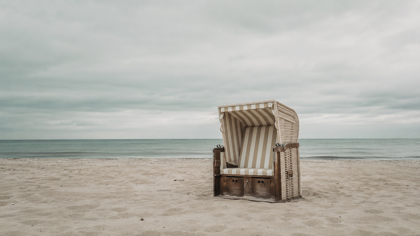 Gestreifter Strandkorb aus Korbgeflecht steht am sandigen Strand unter bewölktem Himmel.